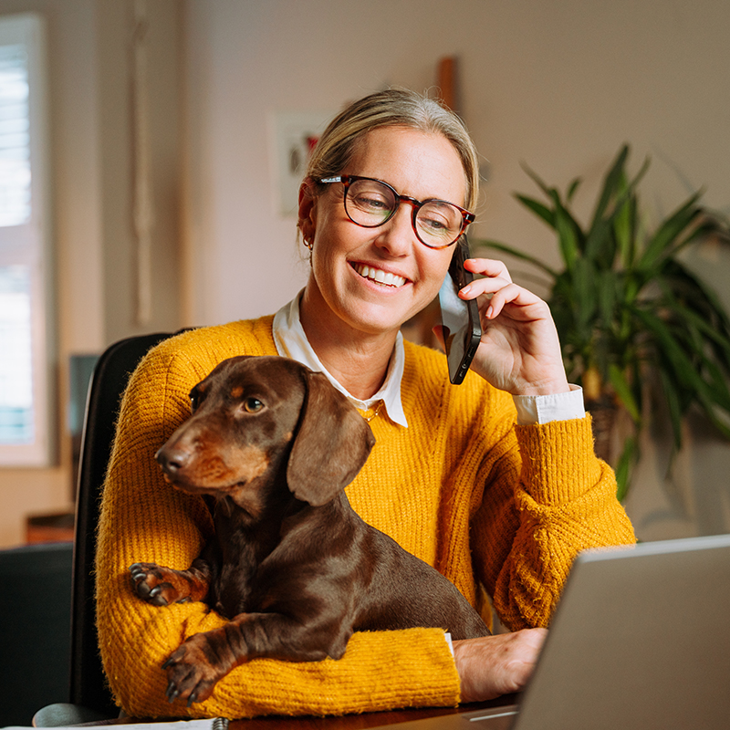 A woman with glasses and smiles while talking on the phone, holding a brown dachshund in her lap.